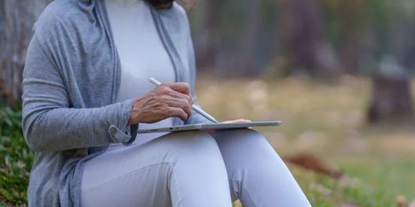 stock image Close up Cheerful overjoyed elderly woman happy and enjoying peaceful beautiful in park and drawing in digital tablet.