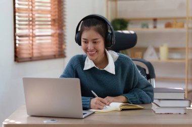 Çevrimiçi eğitim, e-öğrenme. Şık giyinmiş Asyalı bir kadın, dizüstü bilgisayar kullanıyor, online dersler dinliyor, notlar alıyor, evde online çalışmalar yapıyor..