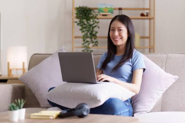 Çekici Asyalı kadın rahat oturma odasında dinleniyor ve dizüstü bilgisayar kullanıyor..