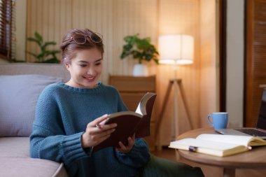 Çekici Asyalı kadın rahat bir oturma odasında dinleniyor ve kitap okuyor, rahatla, kanepe, yaşam tarzı..