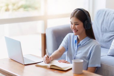 Çevrimiçi eğitim, e-öğrenme. Şık giyinmiş Asyalı bir kadın, dizüstü bilgisayar kullanıyor, online dersler dinliyor, notlar alıyor, evde online çalışmalar yapıyor..