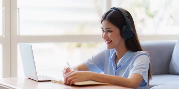 stock image Online education, e-learning. Asian woman in stylish casual clothes, studying using a laptop, listening to online lecture, taking notes, online study at home.