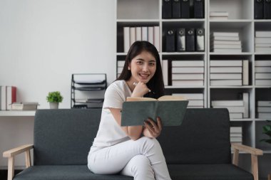 Çekici Asyalı kadın rahat bir oturma odasında dinleniyor ve kitap okuyor, rahatla, kanepe, yaşam tarzı..