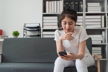 Genç, rahat, gülümseyen, güzel bir kadın evdeki kanepede rahatlıyor. Kadın rahat koltukta cep telefonuyla eğleniyor..