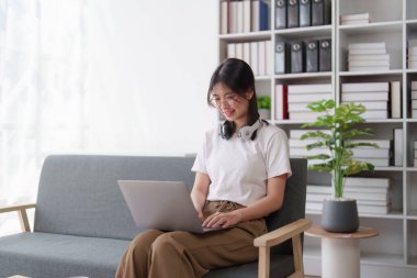 Genç, rahat, gülümseyen, güzel bir kadın evdeki kanepede rahatlıyor. Bir kadın rahat koltukta dizüstü bilgisayarla eğleniyor..