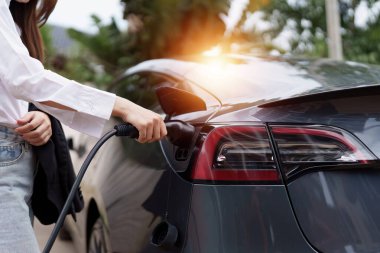 Elektrikli araba şarj oluyor, kadın arabaya elektrik kablosu bağlıyor. Yeşil Enerji kavramı.