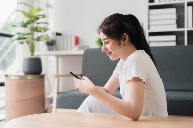 Genç, rahat, gülümseyen, güzel bir kadın evdeki kanepede rahatlıyor. Kadın rahat koltukta cep telefonuyla eğleniyor..