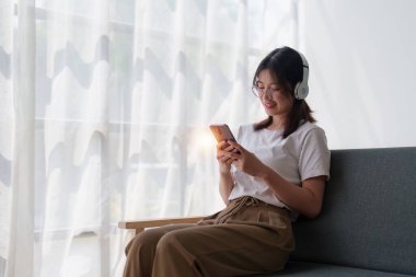 Genç, rahat, gülümseyen, güzel bir kadın evdeki kanepede rahatlıyor. Kadın rahat koltukta cep telefonuyla eğleniyor..