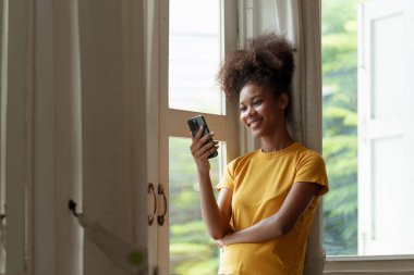 Genç Amerikalı Afrikalı rahat gülümseyen güzel kadın evde dinleniyor. Kadın rahat koltukta cep telefonuyla eğleniyor..