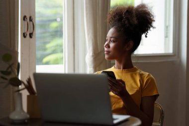 Genç Amerikalı Afrikalı rahat gülümseyen güzel kadın evde dinleniyor. Kadın rahat koltukta cep telefonuyla eğleniyor..