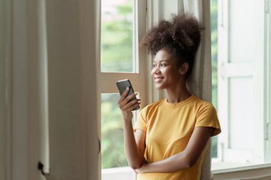 Genç Amerikalı Afrikalı rahat gülümseyen güzel kadın evde dinleniyor. Kadın rahat koltukta cep telefonuyla eğleniyor..