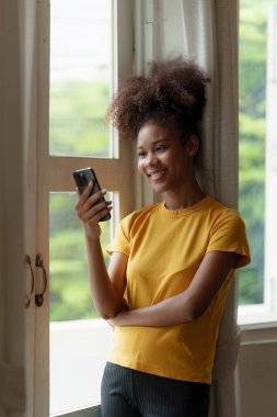 Genç Amerikalı Afrikalı rahat gülümseyen güzel kadın evde dinleniyor. Kadın rahat koltukta cep telefonuyla eğleniyor..