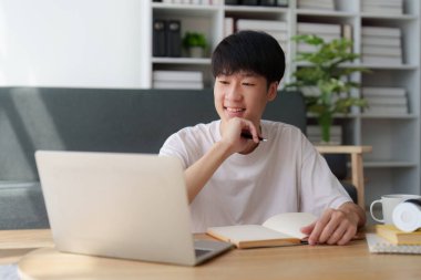 Çevrimiçi eğitim, e-öğrenme. Şık giyinmiş Asyalı bir adam, dizüstü bilgisayar kullanıyor, online dersler dinliyor, notlar alıyor, evde online çalışmalar yapıyor..