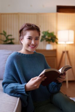 Asyalı bir kadın rahat bir koltukta oturup kitap okuyor..