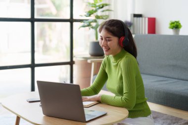 Genç Asyalı rahat gülümseyen güzel kadın evde dizüstü bilgisayarla rahatlıyor. Kadın rahat koltukta cep telefonuyla eğleniyor..