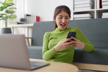 Genç Asyalı rahat gülümseyen güzel kadın evde cep telefonuyla rahatlıyor. Kadın rahat koltukta cep telefonuyla eğleniyor..