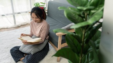 Genç Asyalı kadın evinde kitap okuyor. Oturma odasında yerde oturuyor..
