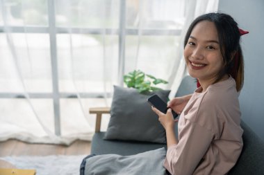 Genç bayan oturma odasında kanepede oturuyor, akıllı telefon kullanıyor ve gülümseyen bir yüzle kameraya bakıyor..