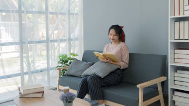 Genç Asyalı kadın evinde kitap okuyor. Oturma odasında, kanepede oturuyor..
