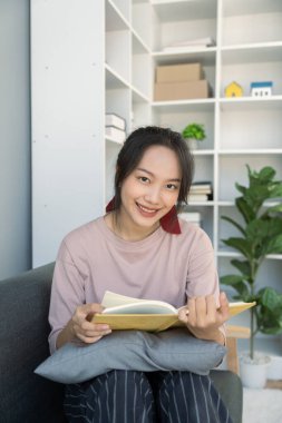 Genç Asyalı kadın evinde kitap okuyor. Oturma odasında, kanepede oturuyor..
