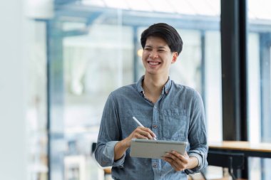 Genç işadamı pencerenin yanında dururken dijital tabletle çevrimiçi çalışıyor..