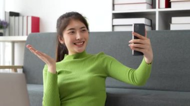 Güzel Asyalı kadın evdeki kanepede oturup arkadaşlarıyla video görüşmesinin keyfini çıkarıyor. Evdeki akıllı telefondan içeriği olan bir kadın. Yüksek kalite 4k görüntü