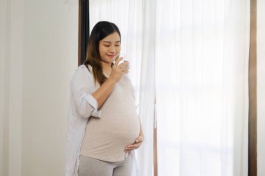 Hamile bir kadın karnına sarılıyor ve pencerede kocaman bir göbekle mutlu oluyor..
