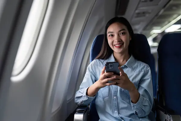 Asian people female person onboard, airplane window, using mobile and looking at camera while on the plane.