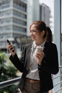 Takım elbiseli genç Asyalı iş kadını şehir caddesinde dikilirken akıllı telefon kullanıyor..