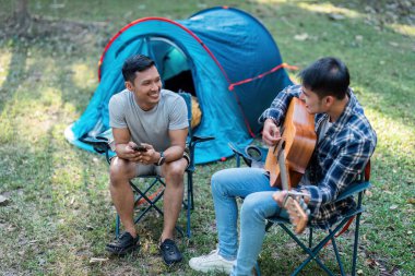 LGBTQIA Gay çifti tatil için ormanda kamp kuruyor ve birlikte gitarda dinleniyorlar..