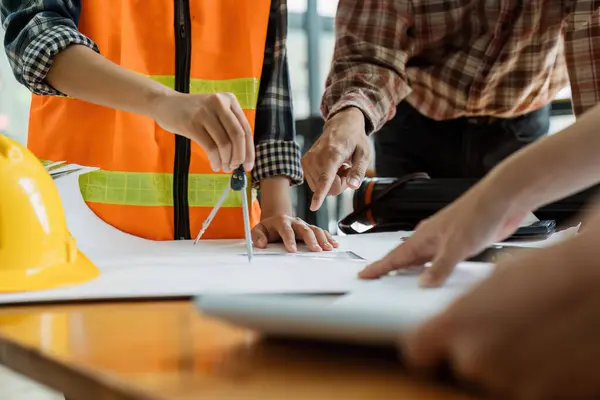 Mühendislerden oluşan bir ekip, ofis masasında mühendislik aletleri bulunan yeni bir inşaat projesinin taslağını inceliyor..