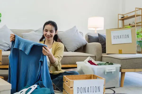 Asian young woman packing clothes at home, putting on stuff into donate box with second hand clothes.