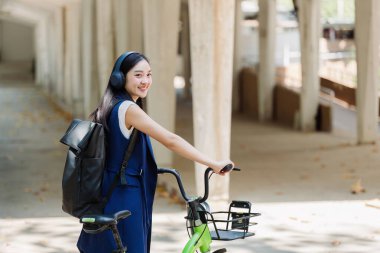 Asyalı genç iş kadını bisikleti çevre dostu yeşil enerji için çalışacak ve şehirde işe giderken kulaklıkla müzik dinleyecek. Çevre dostu.