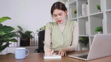 Bir kadın dizüstü bilgisayarla masada otururken bir deftere yazı yazıyor. Yeşil bir yelek giyiyor ve masada bir fincan kahve içiyor.