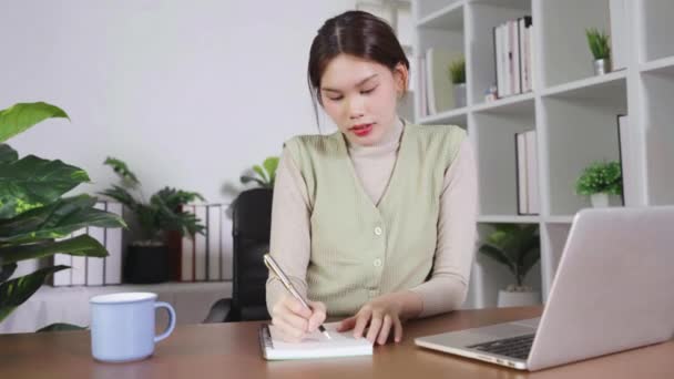 一个女人坐在写字台边 手里拿着笔记本电脑 一边在笔记本上写字 她穿着一件绿色背心 桌上有一杯咖啡 — 图库视频影像