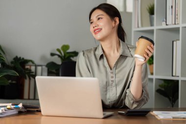 Gülümseyen iş kadını kahve içiyor ve yeşil modern ofiste dizüstü bilgisayarla çalışıyor. Çevre Ofisi güzelmiş. Çevre dostu kavram.