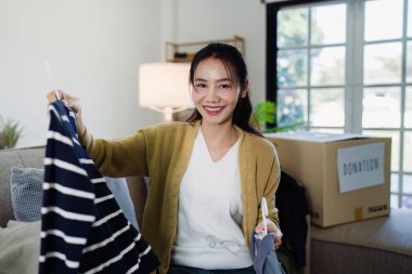 Asyalı bir kadın bağış kutusuna ikinci el kıyafetler koymak için kıyafetlerini evden seçer..