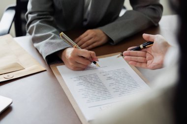 Bir kadın kalemle bir belge imzalıyor. Belge bir dosyanın üzerinde ve kadın bir masada oturuyor.