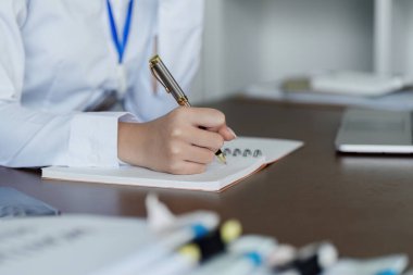 Bir kadın kalemle deftere yazı yazıyor. Beyaz bir gömlek giyiyor ve mavi bir ipliği var. Sahne profesyonel bir ortam öneriyor.