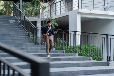 Gülümseyen Asyalı işadamı yeniden kullanılabilir çevre dostu ekolojik fincanı tutuyor ve parkta otururken cep telefonunu kullanıyor..