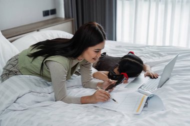 Evden çalışmak, serbest çalışmak ve yaşam tarzı kavramı. Yaratıcı Asyalı kadın portresi dizüstü bilgisayarla yatakta oturuyor ve çalışırken çocuğuna bakıyor..