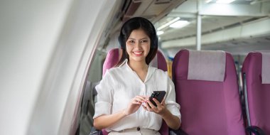 A woman is sitting on a plane with a cell phone in her hand. She is smiling and she is enjoying her time on the plane