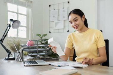 Yurt içi ofis masasında dizüstü bilgisayarla, faturalarla ve belgelerle mali işlerle ilgilenen genç bir kadın, modern bir ev ortamında finansal planlama ve bütçe düzenlemesi kavramı.