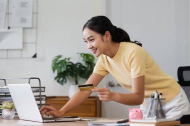 Bilgisayarında online alışveriş için kredi kartı kullanan genç bir kadın ev ofis ortamında modern e-ticaret ve dijital ödeme yöntemleri sergiliyor..