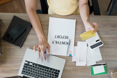 Online ödeme için kredi kartı kullanan bir kadının en üst görüntüsü. Dizüstü bilgisayarı, aylık raporu ve makbuzları olan bir masa..