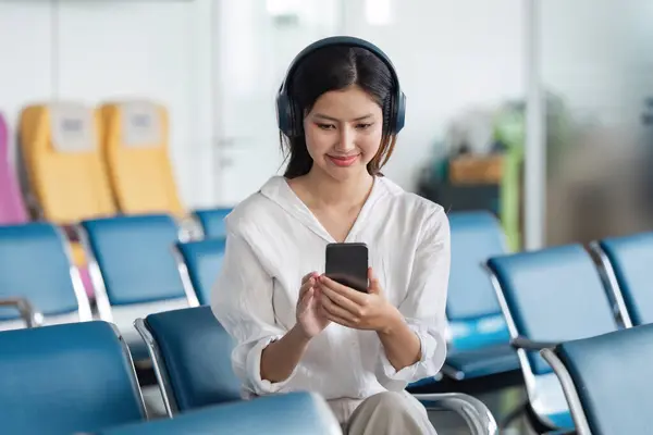 stock image Young woman with headphones using smartphone in airport lounge, ready for travel adventure. Modern air travel experience in comfortable waiting area.