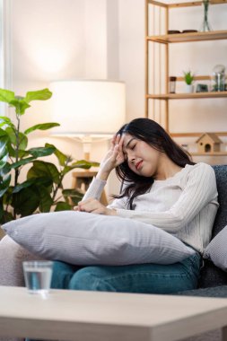 Evde hasta hisseden genç bir kadın, kanepede dinleniyor, eli alnında, rahat ev dekorasyonu ve ev bitkileriyle çevrili..