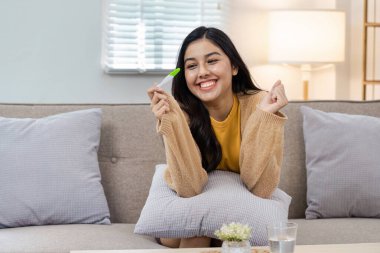 Kanepede oturan neşeli bir kadın, elinde hamilelik testi, olumlu bir sonuç, sıcak bir oturma odasında mutluluğu ifade ediyor..