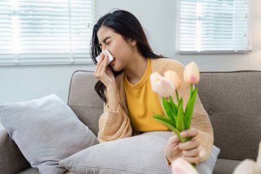 Parlak ve rahat bir oturma odasında elinde bir buket lale tutarken polen alerjisi olan genç bir kadın hapşırıyor..