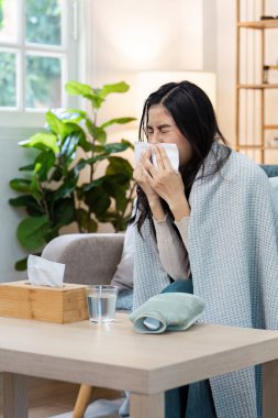 Battaniyeye sarılı bir kadın, sıcak bir oturma odasındaki kanepede oturmuş, soğuk algınlığı konseptini anlatırken mendile hapşırıyor..